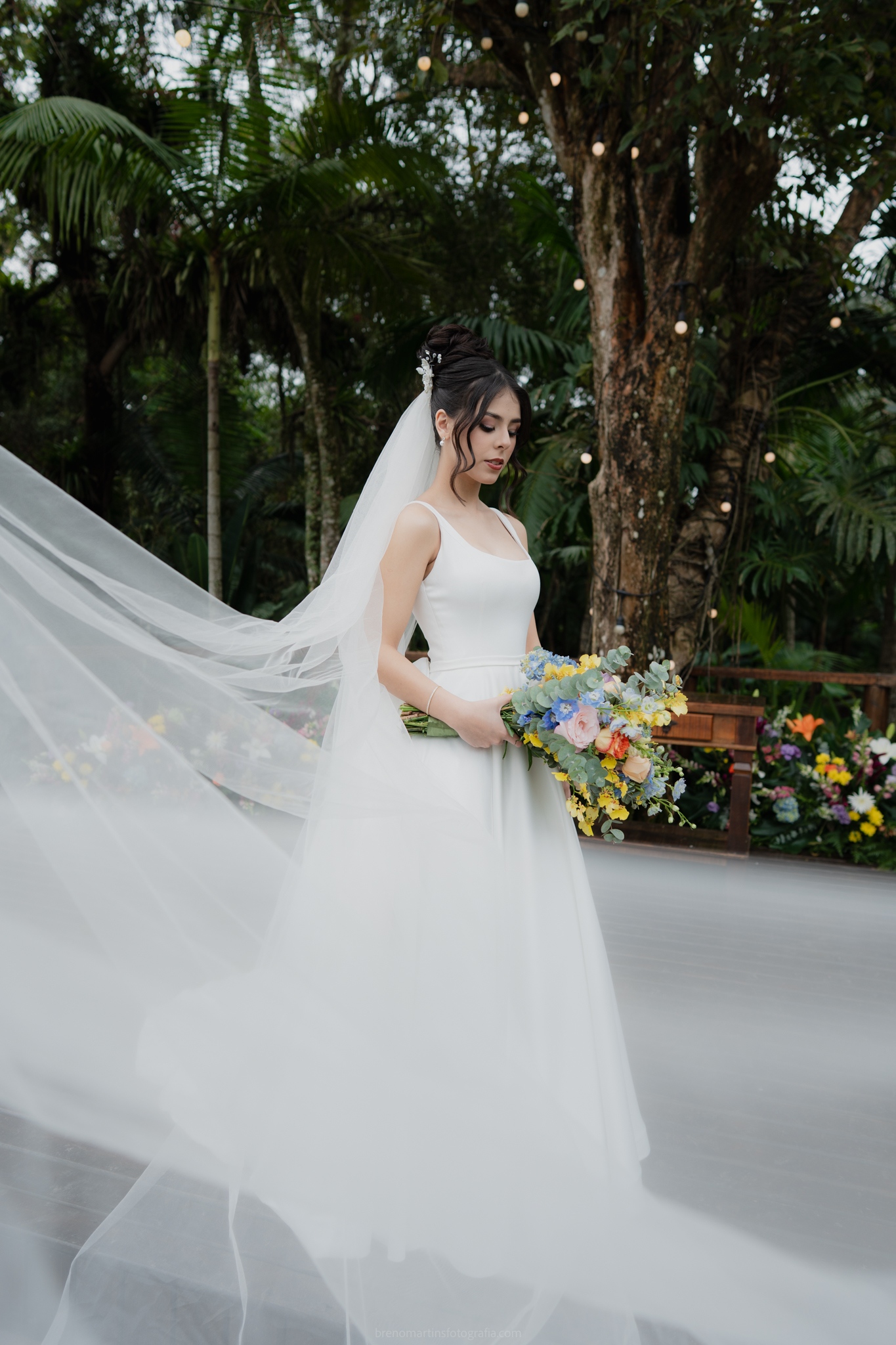 Noiva em vestido branco com véu longo esvoaçante e buquê colorido em um jardim