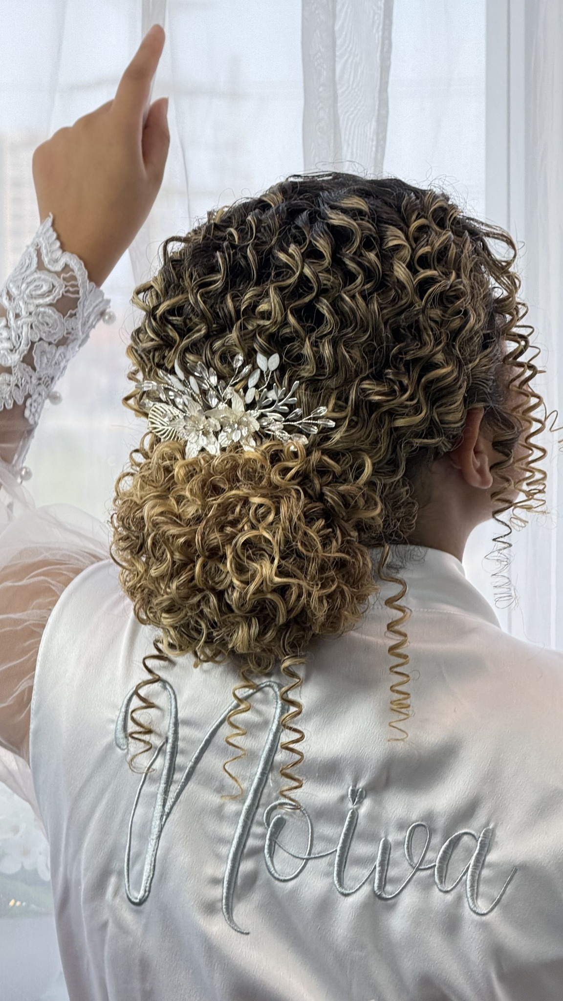 Noiva de costas com coque baixo cacheado e presilha prateada, vestindo robe bordado Noiva
