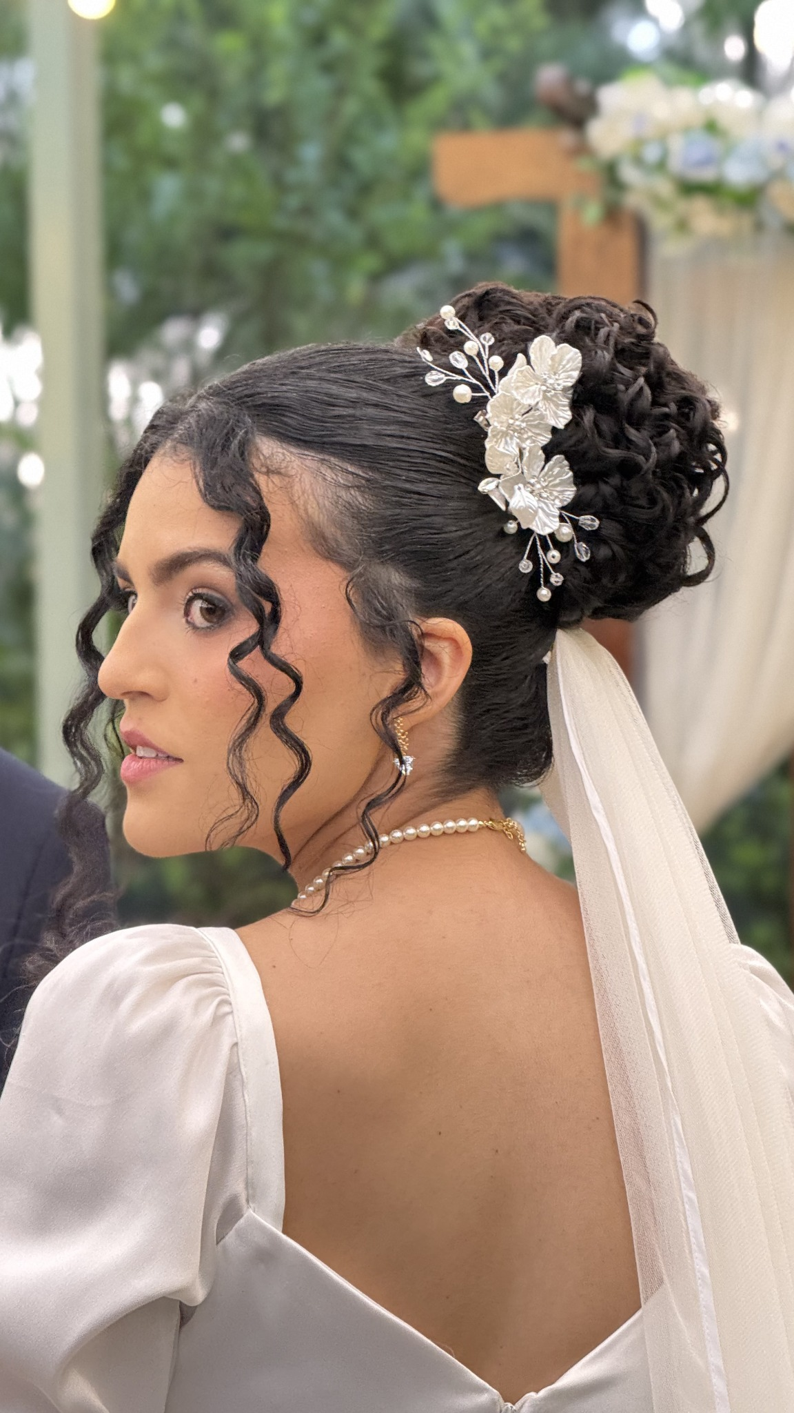 Noiva olhando por cima do ombro com coque alto cacheado e presilha de flores e pérolas