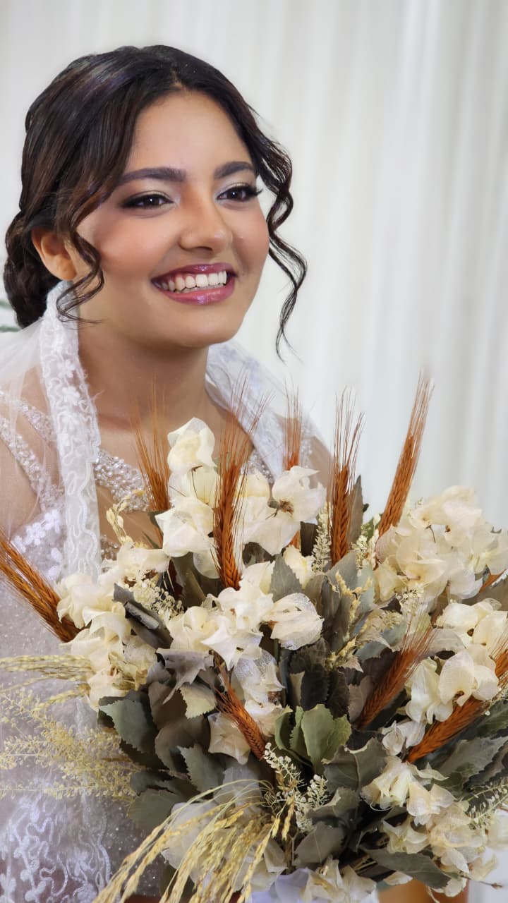 Noiva sorrindo com penteado preso e mechas onduladas, segurando buquê de flores claras e trigo